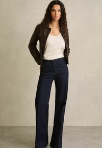 Bruine jas over een witte tanktop, gecombineerd met donkerblauwe wijde jeans. Soepele stof met opvallende stiksels. Neutrale achtergrond.