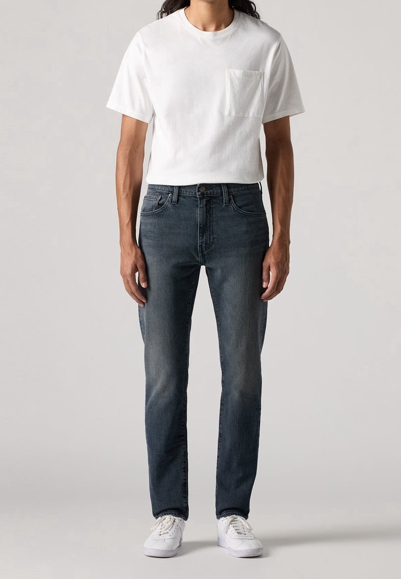 Personne portant un t-shirt blanc à poche, un jean bleu foncé et des baskets blanches, debout devant un fond clair uni.