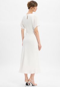 Femme se tenant de profil, portant une robe midi blanche à manches courtes et des sandales à talons hauts argentées, devant un fond blanc uni.