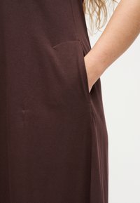 Το χέρι μιας γυναίκας τοποθετημένο στην πλαϊνή τσέπη ενός σκούρου καφέ φορέματος που δείχνει την υφή του υφάσματος και τις λεπτομέρειες της ραφής.