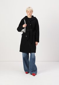 Abrigo negro con cintura ceñida, grandes botones, capucha y mangas largas, combinado con jeans de mezclilla claros acampanados y zapatos rojos.