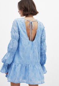 Svetlo modra obleka s cvetličnim vezenjem, dolgimi puhastimi rokavi, odprtim hrbtom in zaveznim detajlom. Ima večplastni volanček in teksturirano tkanino.
