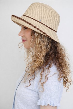 Woman with curly blonde hair wearing a beige straw sun hat and light blue short-sleeve textured top, facing left against a plain background.