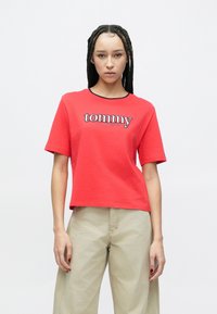 Hosszú fonott hajú nő piros, rövid�ített "tommy jeans" feliratú pólóban és bézs, bőszárú nadrágban, fehér háttér előtt állva.