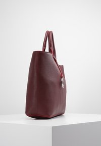 Burgundowa skórzana torba tote o gładkiej fakturze, z podwójnymi uchwytami, czerwonymi akcentami i srebrnym zawieszką przypiętą do suwaka.