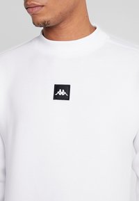 Uomo che indossa un felpa bianca con un piccolo logo quadrato nero che presenta due silhouette specchiate sedute sul petto.