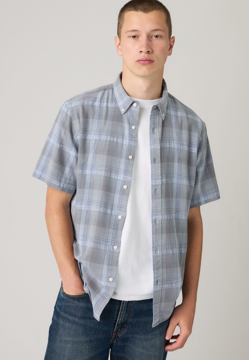Jeune homme portant une chemise à manches courtes à carreaux bleu-gris par-dessus un t-shirt blanc avec un jean bleu, debout avec une main dans la poche.