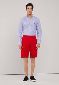 Pantalones cortos rojos y una camisa de botón a rayas blanca y azul. La camisa tiene un pequeño logo en el pecho. Se lleva con zapatillas oscuras.