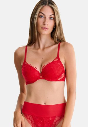 Femme aux longs cheveux blonds portant un soutien-gorge en dentelle rouge et une culotte taille haute assortie, debout devant un fond blanc uni.