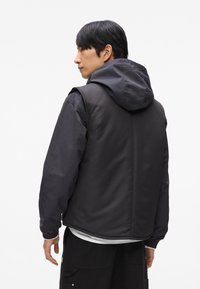 Melna pūkāmā jakas ar kapuci, ar gludu tekstūru, elastīgiem piedurkņu galiem un slāņotu dizainu. Aizmugurē ir vienkārša, tīra apdare.