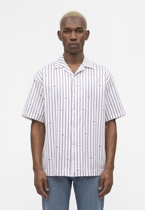 Άνδρας με κοντά ξανθά μαλλιά που φοράει λευκό κοντομάνικο πουκάμισο με κάθετες ρίγες και κόκκινα διαμάντια.