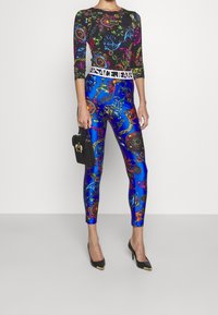Top negro de manga larga con patrones multicolores, combinado con leggins azules con diseños ornamentales, usado con tacones negros y un bolso pequeño.