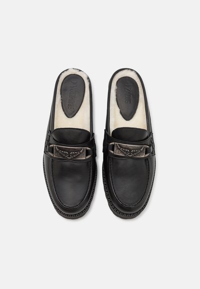 Black leather slip-on shoes featuring a fur-lined interior, decorative metallic buckle with an embossed design, and contrasting stitching.