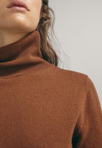 Gros plan d'une personne portant un pull col roulé côtelé marron, montrant le cou, l'épaule et le bas du visage avec des mèches de cheveux lâches.