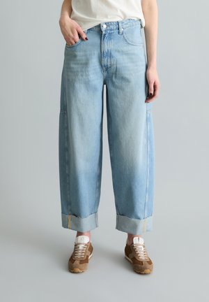 Person trägt hellblaue Jeans mit weitem Bein und hochgekrempelten Säumen, weißes Oberteil und braune Sneakers, steht vor einem einfarbigen grauen Hintergrund.