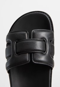 Sandale à glissière en cuir noir avec une large bride ornée d'une boucle décorative. Texture lisse et semelle intérieure moulée. Design à semelle plate.