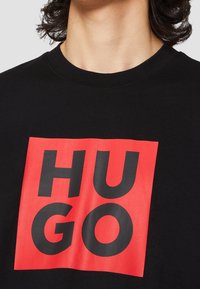 Czarny bawełniany t-shirt z dużą czerwoną grafiką w kształcie kwadratu oraz wyraźnym czarnym napisem "HUGO" na klatce piersiowej. Klasyczny krój z okrągłym dekoltem.