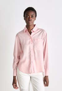 Camicia button-up a maniche lunghe di un rosa chiaro, con colletto e taschino. Il tessuto appare liscio e leggermente aderente, con una silhouette rilassata.
