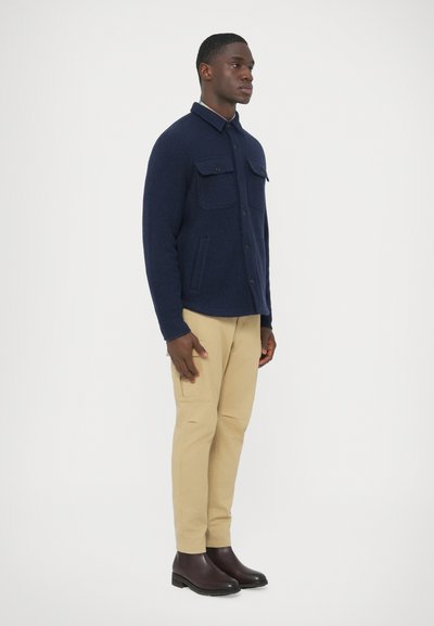 Casaco azul-marinho em mistura de lã com dois bolsos no peito, fecho de botão e colarinho, combinado com calças bege e botas pretas até ao tornozelo.