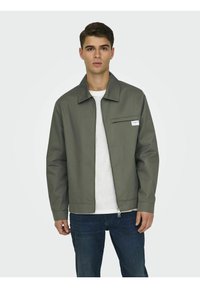 Chaqueta verde oliva con cremallera, cuello puntiagudo, bolsillo en el pecho y puños elásticos, combinada con una camisa blanca y jeans azules.