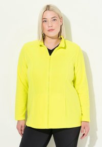 Kirkkaan keltainen fleece-takki, jossa on vetoketjukiinnitys, korkea kaulus ja sivutaskut. Pehmeä kangas ja rakenteellinen istuvuus. Musta toppi alla.