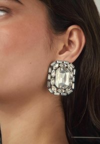 Ohr einer Frau, die einen großen, rechteckigen Statement-Ohrring trägt, besetzt mit verschiedenen klaren, geschliffenen Kristallen in einer silbernen Fassung.