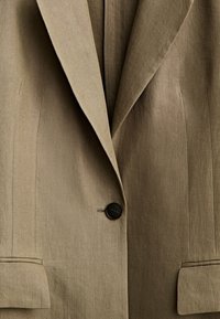 Vaaleanruskea bleiseri, jossa on teksturoitu kangas. Varustettu notched-lapelsilla, kahdella etutaskulla ja yhdellä mustalla nappikiinnityksellä.