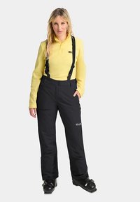 Schwarze wasserdichte Skihose mit verstellbaren Schulterriemen, kombiniert mit einem leichten gelben Langarmshirt, getragen mit schwarzen Skischuhen.