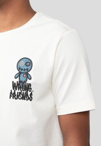 Camiseta blanca de algodón con un personaje de dibujos animados azul, con ojos de botón y detalles cosidos, acompañada de un texto en negrita que dice "AMIGOS INCORRECTOS".