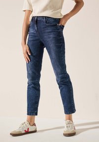 Jean en denim bleu marine avec une coupe slim et un design à cinq poches, présentant une fermeture éclair et un minimalisme dans les détails. Associé à des baskets blanches.