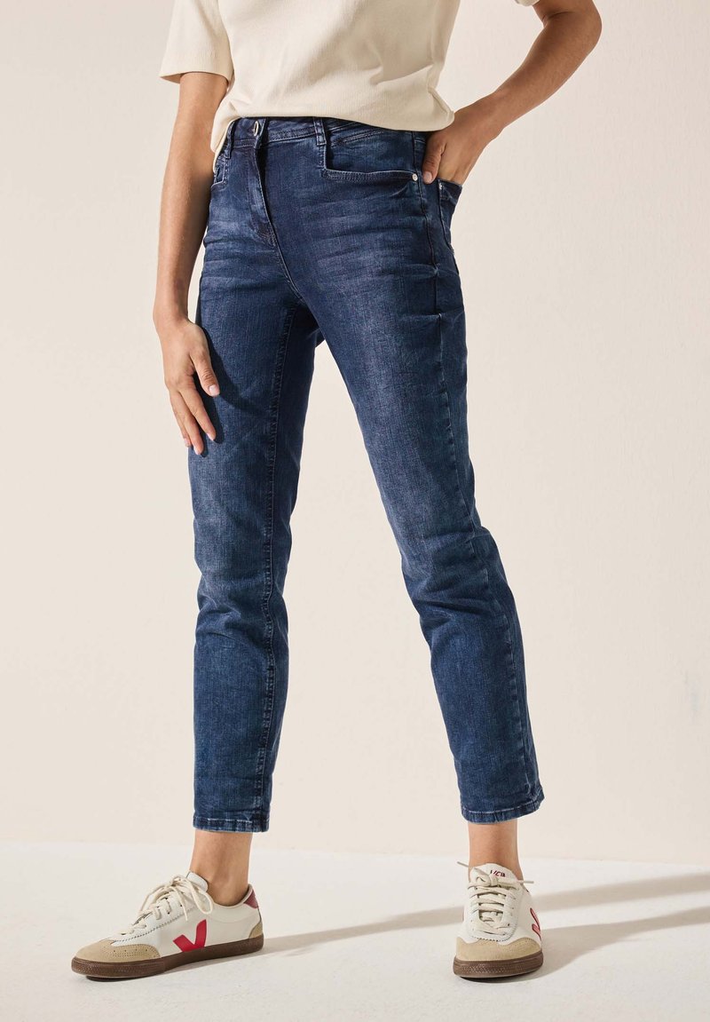 Jean en denim bleu marine avec une coupe slim et un design à cinq poches, présentant une fermeture éclair et un minimalisme dans les détails. Associé à des baskets blanches.