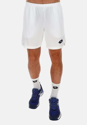 Uomo con pantaloncini sportivi bianchi, calzini bianchi alla caviglia e scarpe sportive blu, in piedi contro uno sfondo semplice.