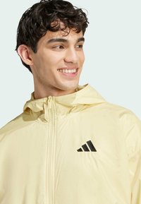 Lys gul vindjakke med lynlås foran, hætte og sort Adidas-logo. Glat tekstur og letvægtsdesign.