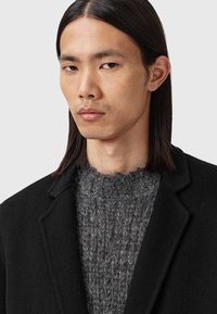 Cămașă neagră din lână peste un pulover texturat de culoare gri, evidențiind un design structurat și linii curate. Finisaj neted cu guler ajustat.