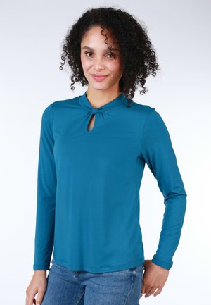 Teal long-sleeve top with a keyhole neckline and knotted detail at the collar. Soft fabric with a smooth texture, paired with casual jeans.