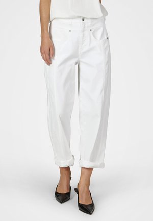 Person trägt weiße, hochgekrempelte Jeans und schwarze spitze High Heels, steht vor einem schlichten hellen Hintergrund.