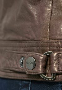 Bruine leren jackedetail met een gestructureerd oppervlak, decoratieve stiksels en een metalen knop met gegraveerde markeringen aan de zijkant.