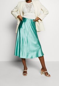 Kvinna iklädd en krämfärgad blazer, vit spetsblus, teal satin midikjol och krämfärgade högklackade sandaler, poserar med händerna på höfterna.