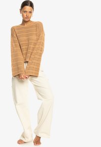 Tan knit sweater with wide sleeves and open weave pattern, paired with loose-fitting white trousers. Natural fabric texture visible.