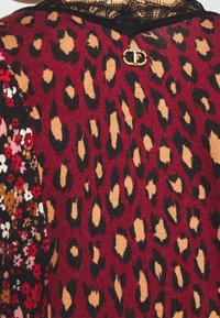 Leopardikuosinen pusero, jossa viininpunainen tausta sekä mustia ja ruskeita laikkuja. Musta pitsikaulus ja kukkakuosi toisella puolella. Kullanväriset yksityiskohdat.