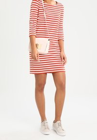 Czerwona i biała sukienka w paski z rękawami 3/4, dekoltem w łódkę, w połączeniu z lekką torebką na ramię i beżowymi sneakersami.