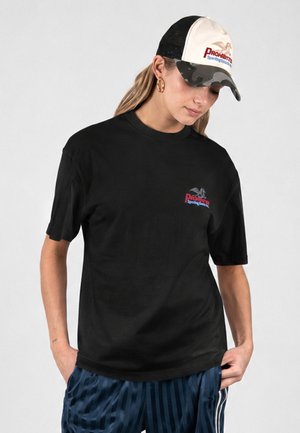 Kvinde iført sort T-shirt med print og blå stribede bukser samt en hvid og camouflagefarvet baseballcap, stående med hænderne i lommerne.