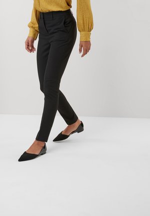 Person iført stramme sorte bukser, sorte spidse flade sko og en langærmet bluse i sennepsgul, gående foran en ensfarvet hvid baggrund.