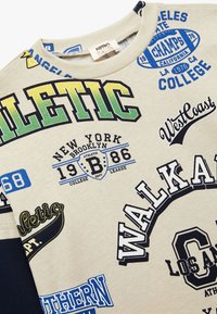Cremefarbener Sweatshirt mit verschiedenen bedruckten Patches und Texten in Blau, Grün, Gelb und Schwarz. Rippenkragen; aus Baumwollmaterial.