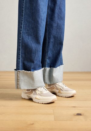 Persoon in wijdvallende, opgerolde blauwe jeans en beige veterschoenen staat op een houten vloer.