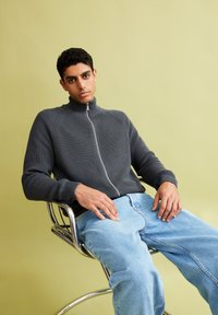 Grauer gerippter Reißverschluss-Pullover mit hohem Kragen, kombiniert mit hellblauen, locker sitzenden Jeans, sitzend auf einem Chromstuhl vor einem hellgrünen Hintergrund.