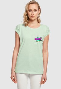 Mintgrøn kortærmet t-shirt lavet af blødt stof, med et farverigt "BOOM!" grafik på venstre brystområde. Hvide bukser synlige.