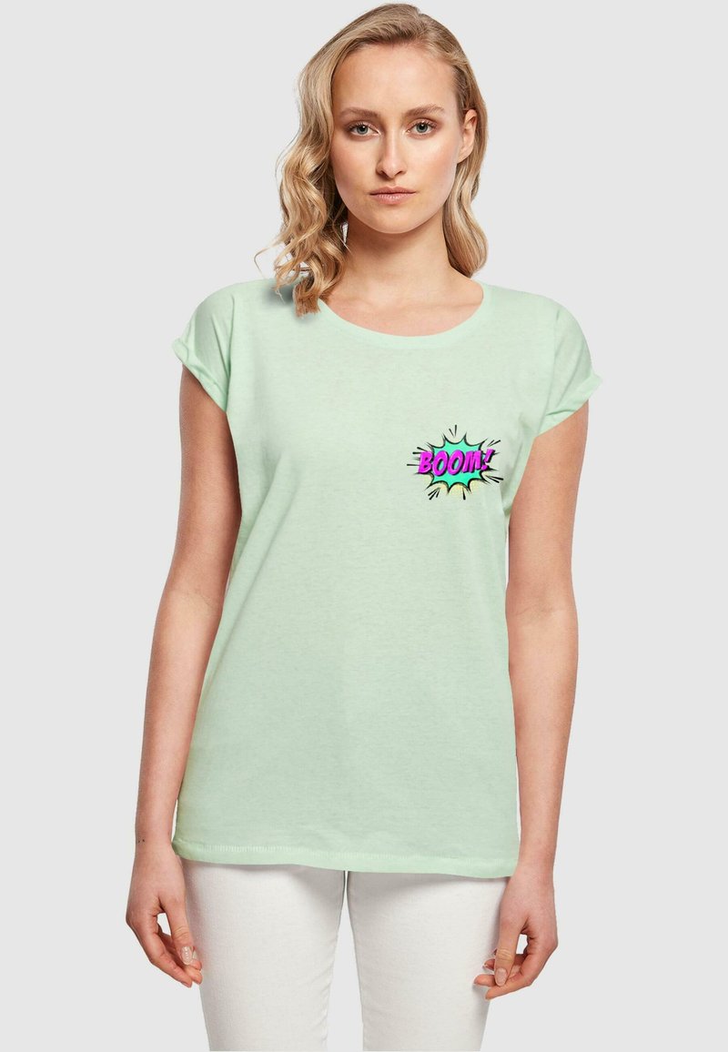 Mintgrøn kortærmet t-shirt lavet af blødt stof, med et farverigt "BOOM!" grafik på venstre brystområde. Hvide bukser synlige.