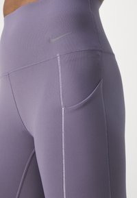 Violet sportiniai antblauzdžiai su pritaikyta forma, turintys lygų paviršių, šonines kišenes ir mažą pilką Nike logotipą ant juosmens.