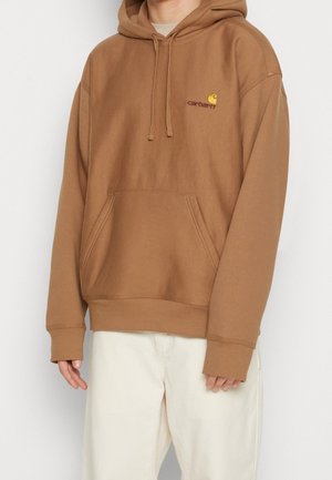 Person wearing a brown Carhartt hoodie with front pocket and drawstrings, paired with white pants, against a plain white background.